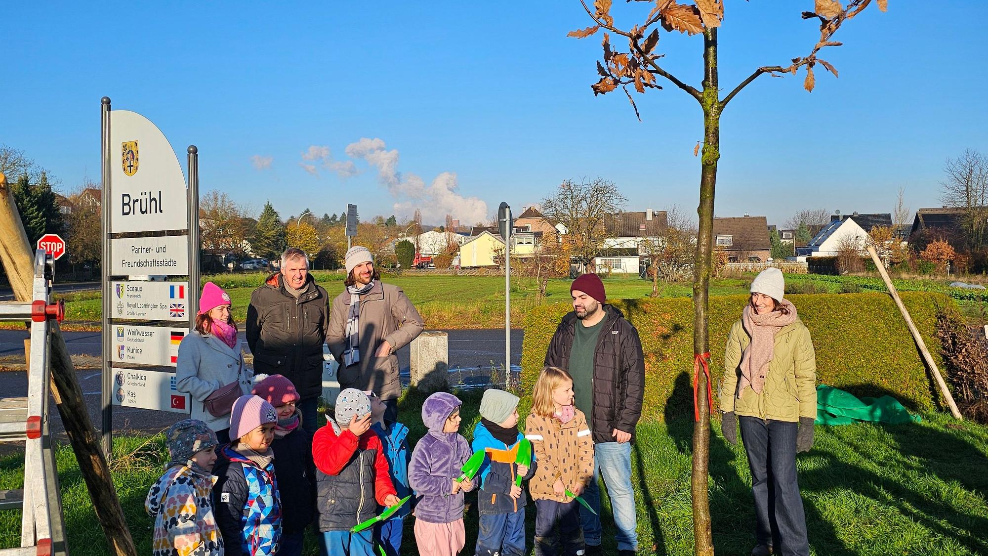 Kinder und Erwachsene stehen neben einem neu gepflanzten Baum.
