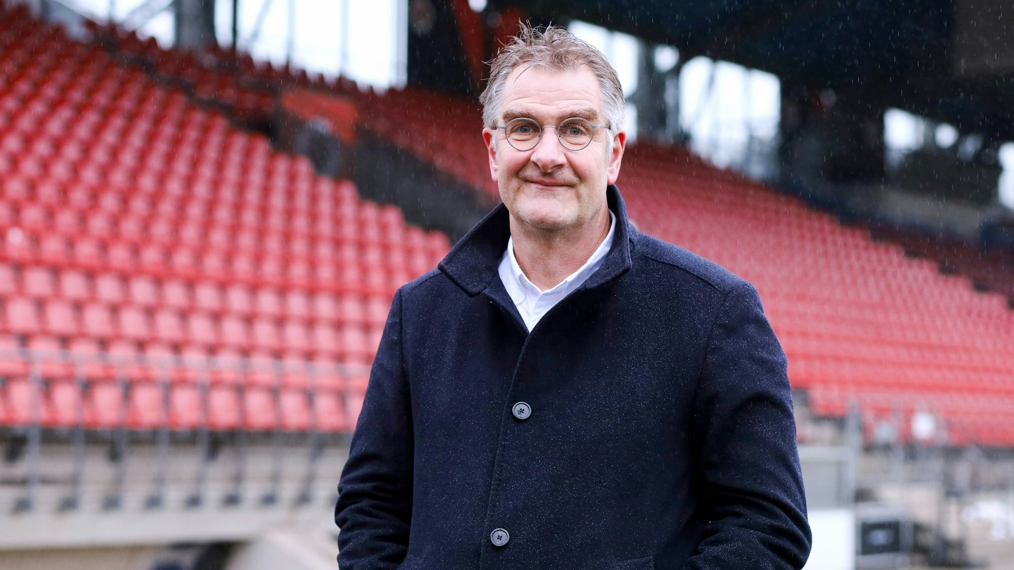 Jürgen Wiebicke steht vor einer Tribüne mit roten Sitzen.