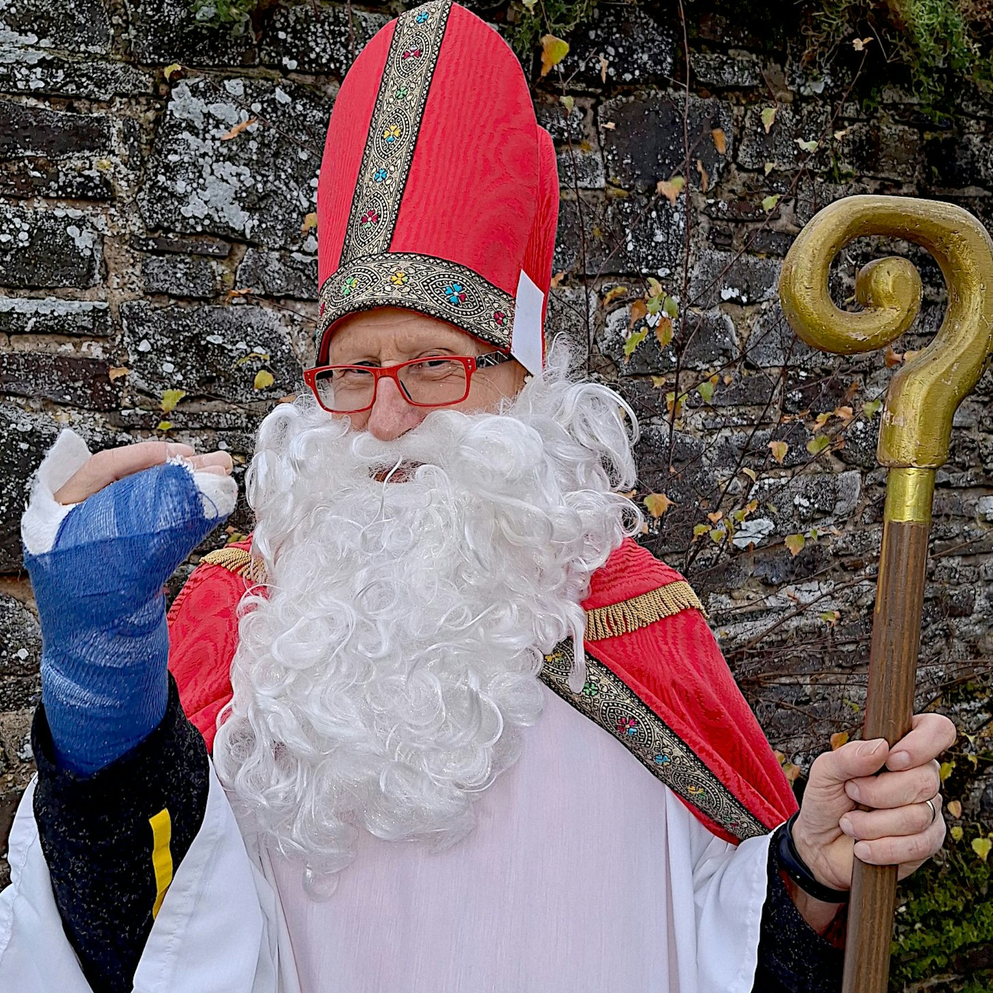 Ein Mann im Nikolauskostüm hält seine rechte Hand hoch, an der er eine blau umwickelte Schiene trägt.