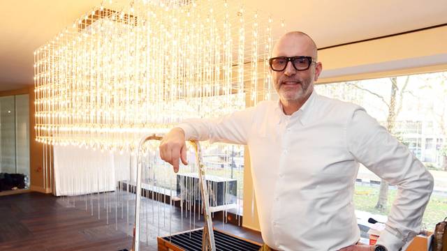 Ein Mann in weißem Hemd und mit Brille steht in seinem Laden vor einem Glasleuchter.