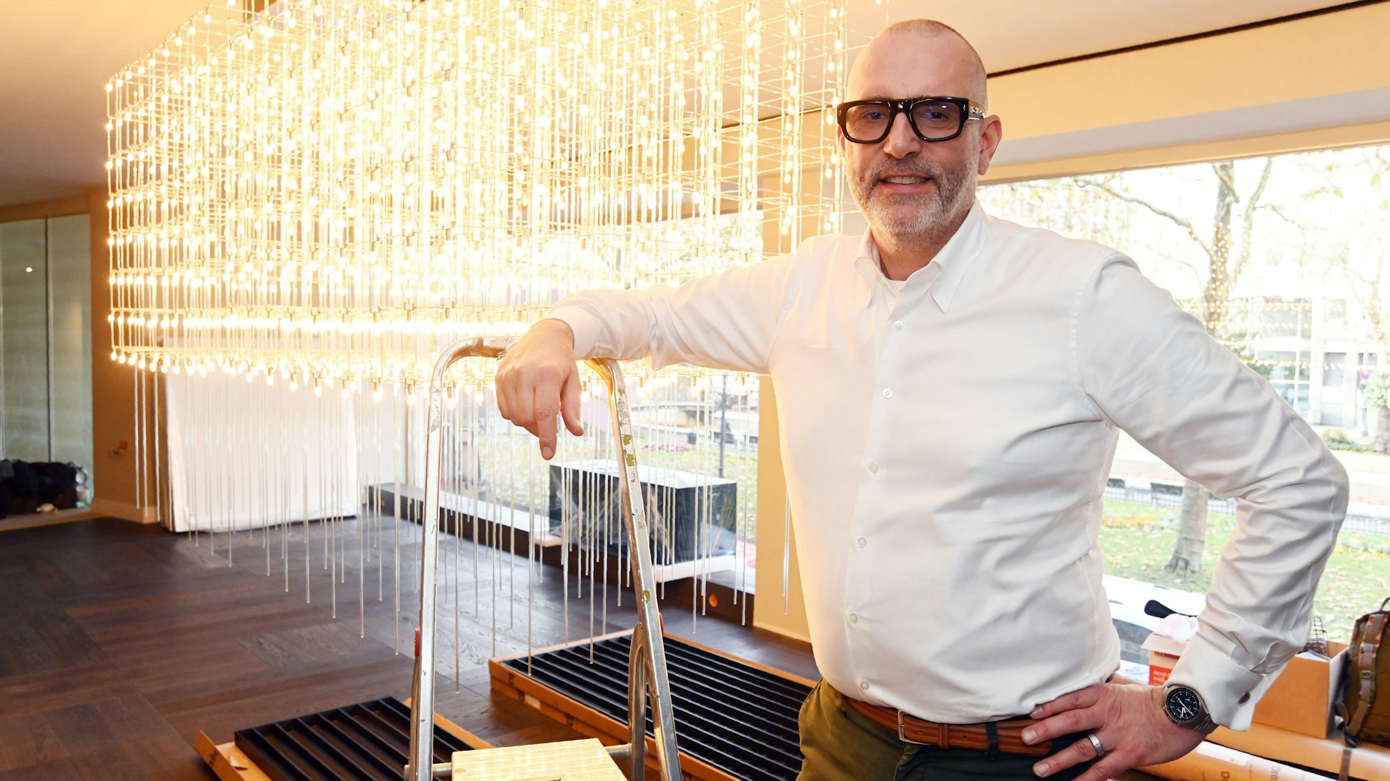 Ein Mann in weißem Hemd und mit Brille steht in seinem Laden vor einem Glasleuchter.