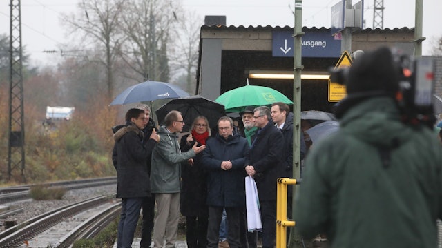 Mehrere Männer und eine Frau auf einem Bahnsteig, im Vordergrund ein Mann mit einer Kamera.