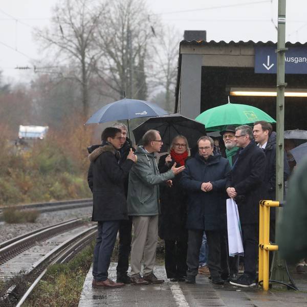 Mehrere Männer und eine Frau auf einem Bahnsteig, im Vordergrund ein Mann mit einer Kamera.