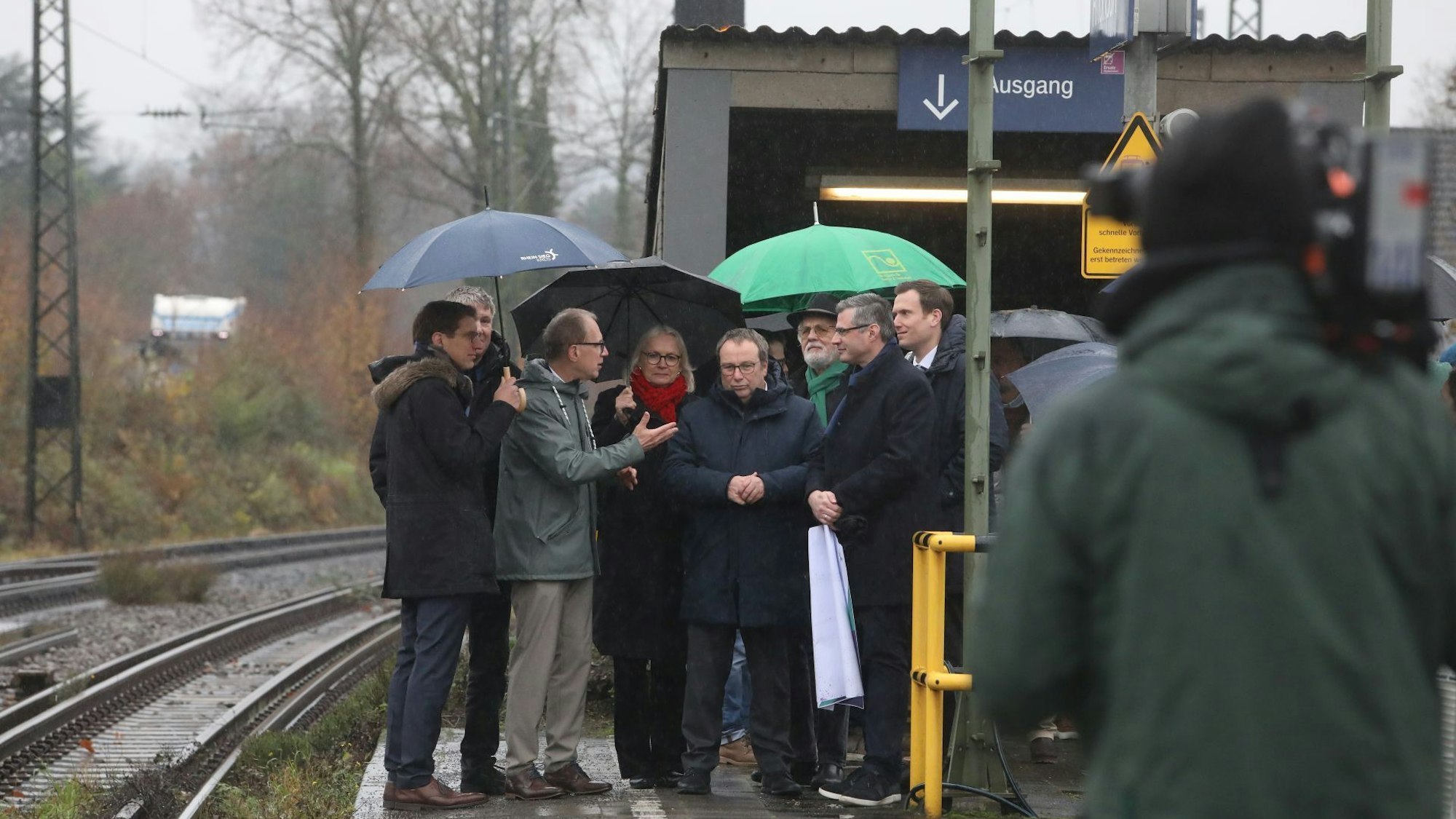 Mehrere Männer und eine Frau auf einem Bahnsteig, im Vordergrund ein Mann mit einer Kamera.