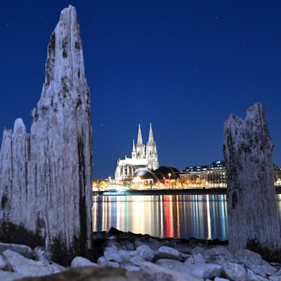 Der Kölner Dom wird am Abend durch Licht erhellt. Teile einer Holzkonstruktion, die normalerweise unter Wasser liegen, sind am Rhein zu sehen.
