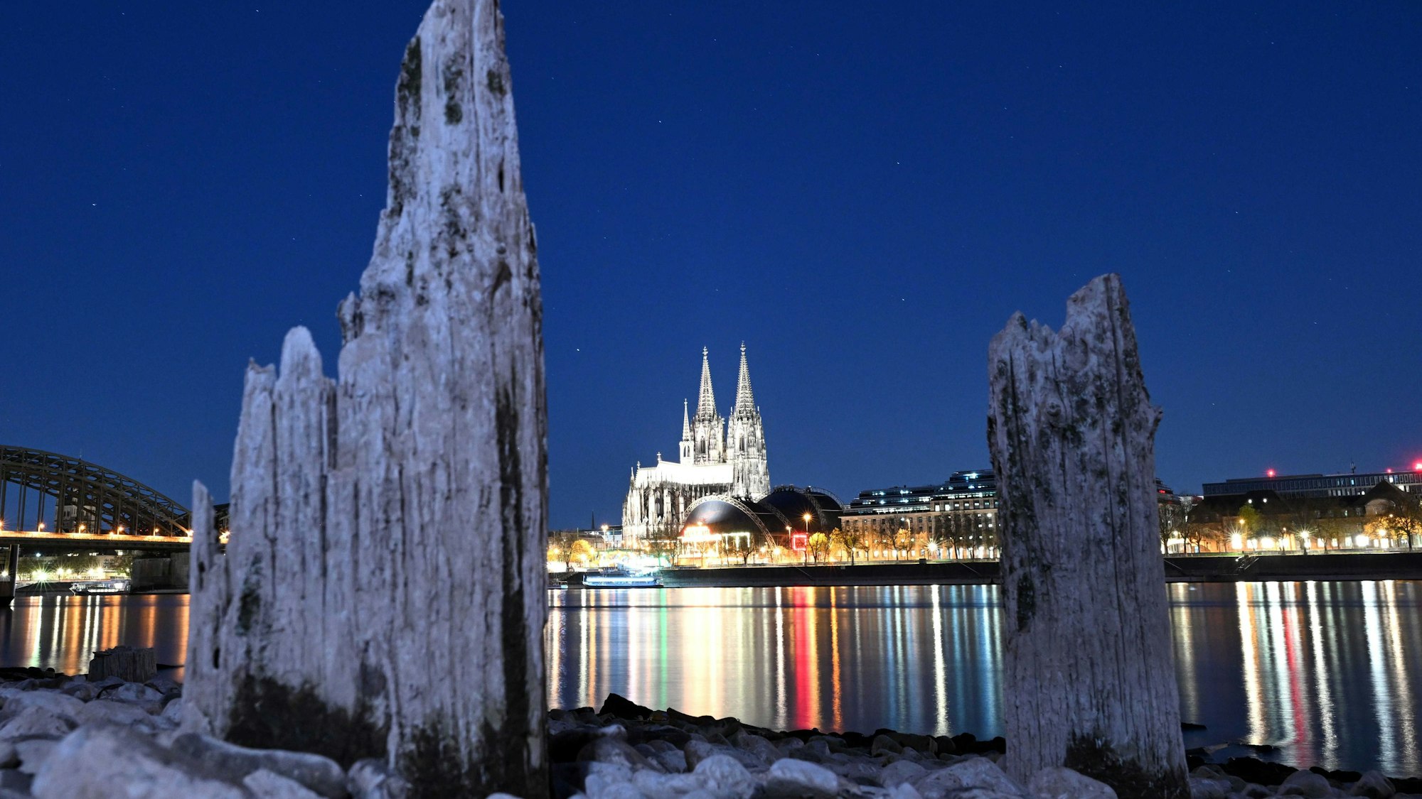 Der Kölner Dom wird am Abend durch Licht erhellt. Teile einer Holzkonstruktion, die normalerweise unter Wasser liegen, sind am Rhein zu sehen.