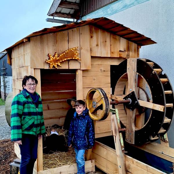 Eine Frau und ihr Enkelsohn stehen an einer gezimmerten Wassermühle, in der eine Krippe für den Krippenweg in Blankenheim-Ripsdorf eingerichtet ist.