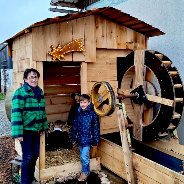 Eine Frau und ihr Enkelsohn stehen an einer gezimmerten Wassermühle, in der eine Krippe für den Krippenweg in Blankenheim-Ripsdorf eingerichtet ist.