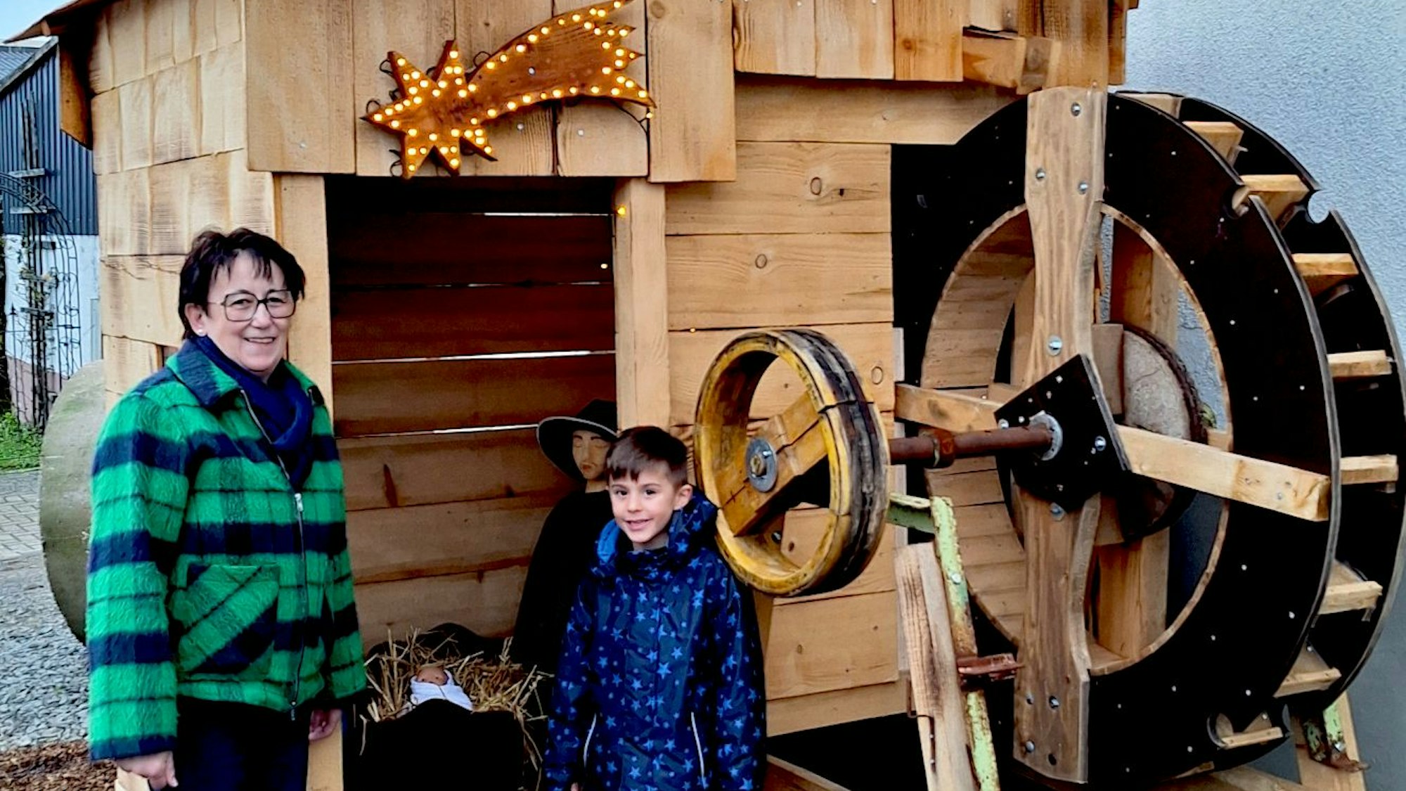 Eine Frau und ihr Enkelsohn stehen an einer gezimmerten Wassermühle, in der eine Krippe für den Krippenweg in Blankenheim-Ripsdorf eingerichtet ist.