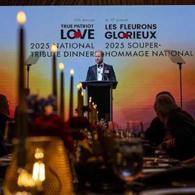 Prinz Harry spricht bei einem Gala-Dinner vor einer großen Leinwand mit zweisprachiger Aufschrift.