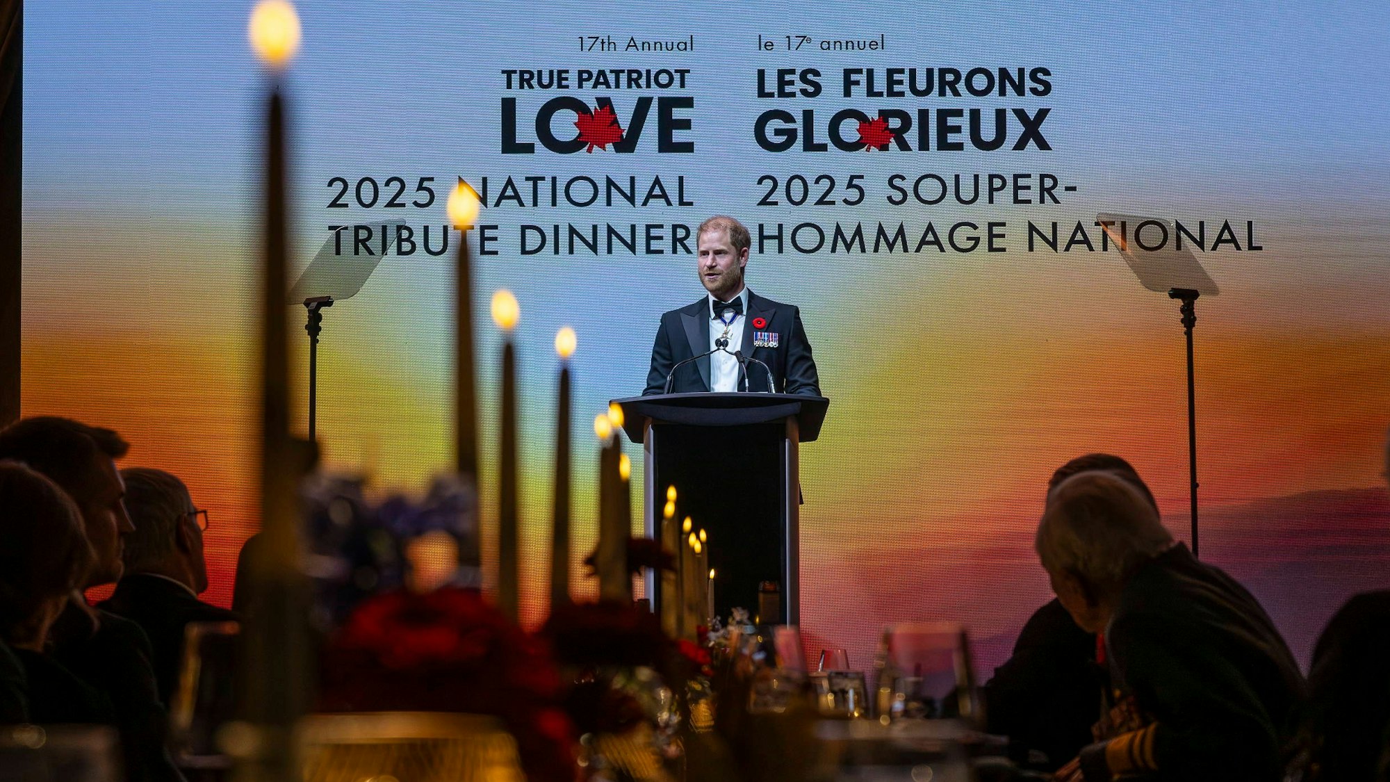 Prinz Harry spricht bei einem Gala-Dinner vor einer großen Leinwand mit zweisprachiger Aufschrift.