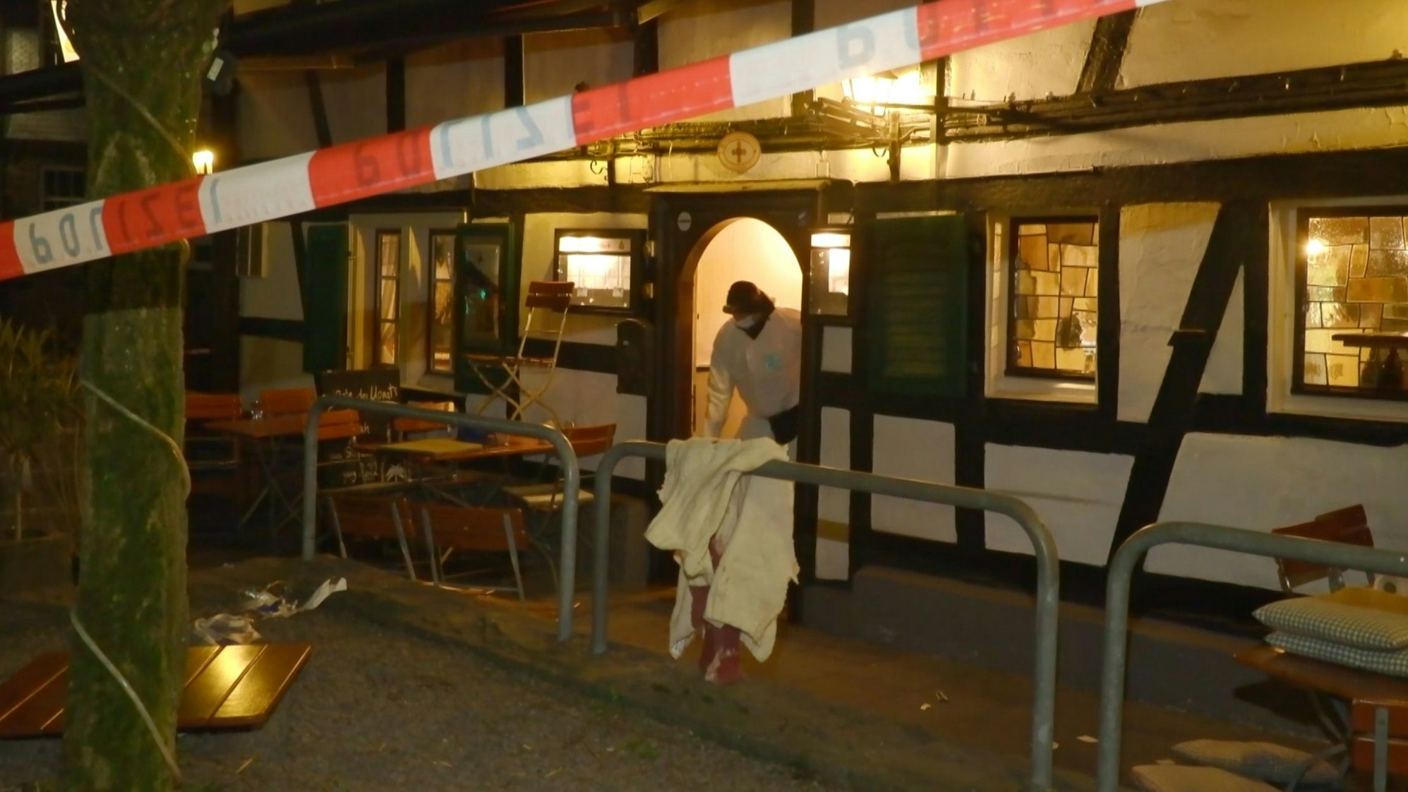Ein Polizeiflatterband sperrt eine Gaststätte in Rösrath-Hoffnungsthal am Abend des 17. März 2025 ab. Ein Ermittler der Polizei steht in der Tür.