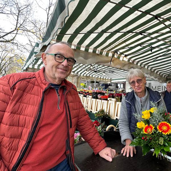 Ein Mann in roter Jacke steht an einem Blumenstand. Die Verkäuferin reicht ihm einen Strauß.