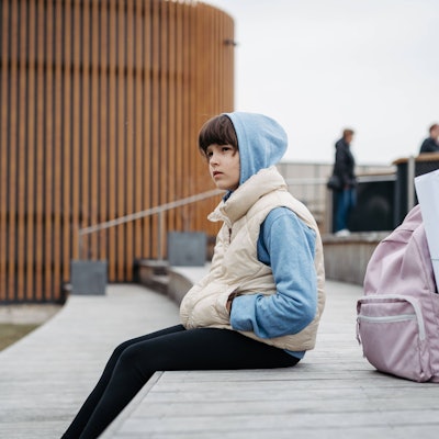 Ein Mädchen sitzt mit ihrer Schultasche und hochgezogener Kapuze auf einem Schulhof.