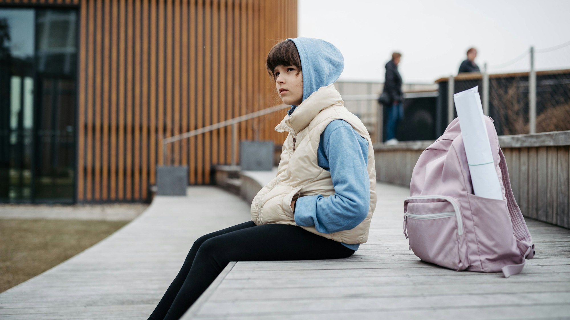 Ein Mädchen sitzt mit ihrer Schultasche und hochgezogener Kapuze auf einem Schulhof.