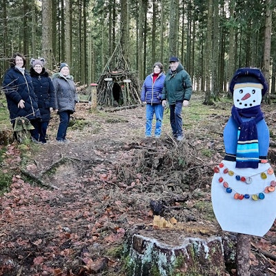 Mitten im Wald stehen die Personen, die den Krippenweg vorgestellt haben. Im Hintergrund sieht man die Schutzhütte, im Vordergrund einen selbstgebastelten Papp-Schneemann mit Schal.