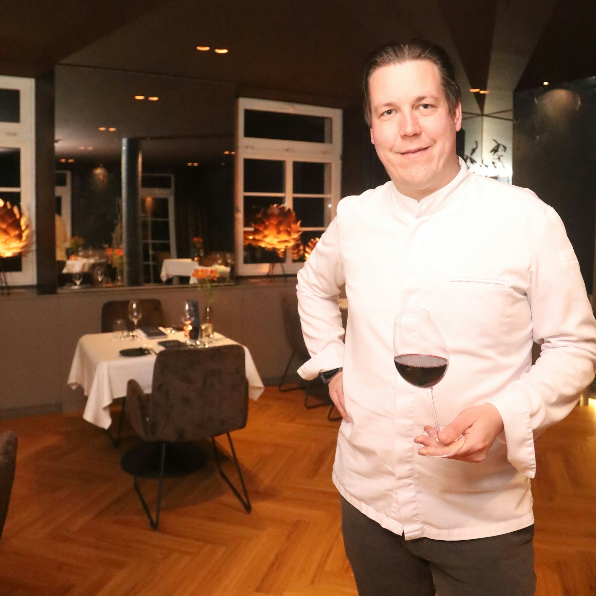 Thomas Gilles steht in Kochjacke im Restaurant und hält ein Glas Rotwein in der Hand. Er lächelt in die Kamera.