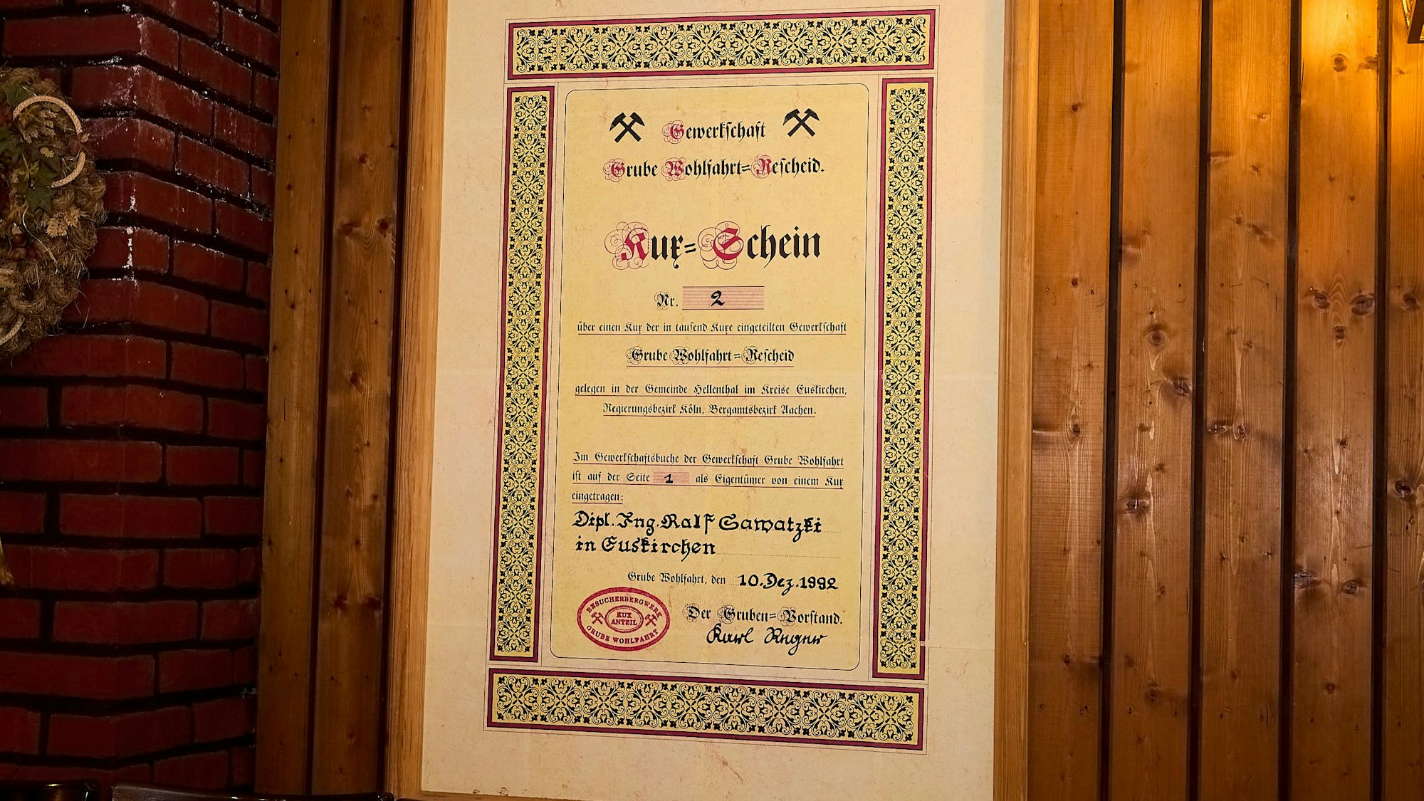 Das Bild zeigt einen gerahmten Kux, der an einer Holzwand hängt.