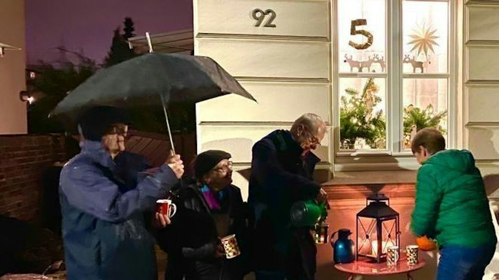 Männer und Frauen vor einem weihnachtlich geschmückten Fenster; auf einem Tisch stehen eine Laterne, Tassen und Thermoskanne.