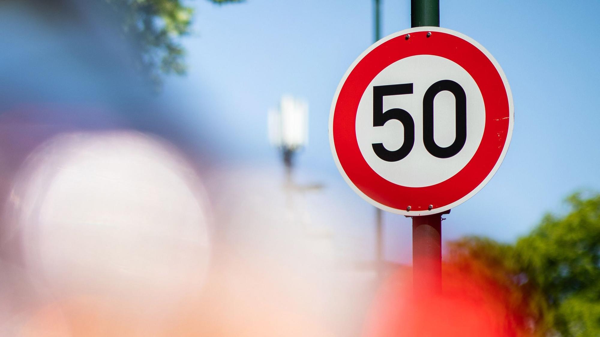 Ein Verkehrsschild zur Geschwindigkeitsbegrenzung auf Tempo 50 steht auf einer Hauptstraße vor geparkten Autos.