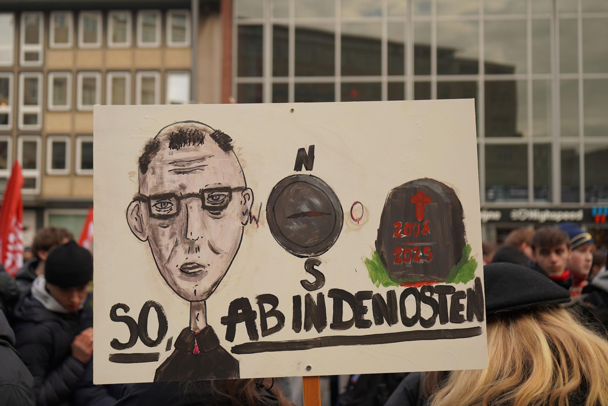 Eine Karikatur von Friedrich Merz, die Aufschrift „Ab in den Osten“ und ein Grabstein sind auf dem Schild zu sehen.