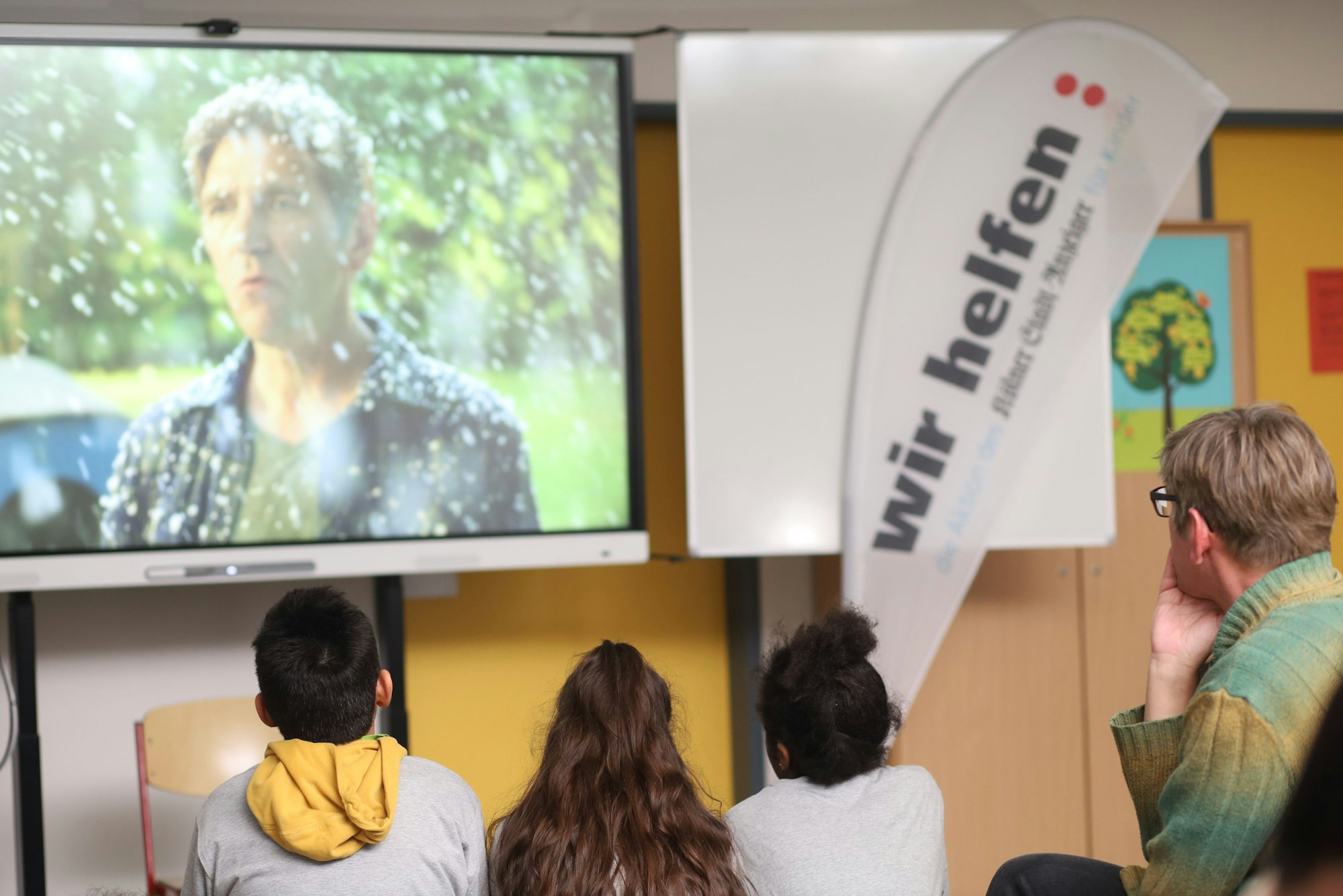 Drei junge Mitglieder der Linoclub-Kinderrats sitzen gemeinsam mit Fritz-Fuchs-Darsteller Guido Hammesfahr vor einem großen TV-Bildschirm