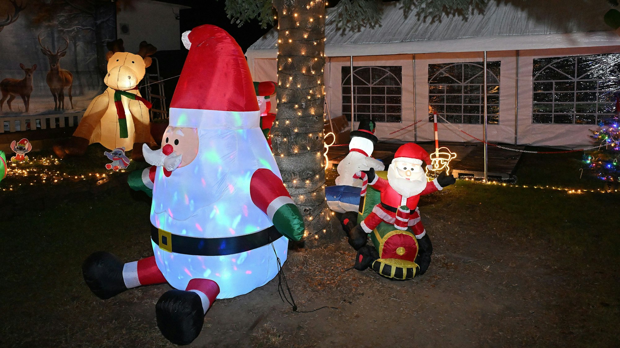 Ein großer aufblasbarer Weihnachtsmann sitzt vor einem Baum mit Lichterkette.