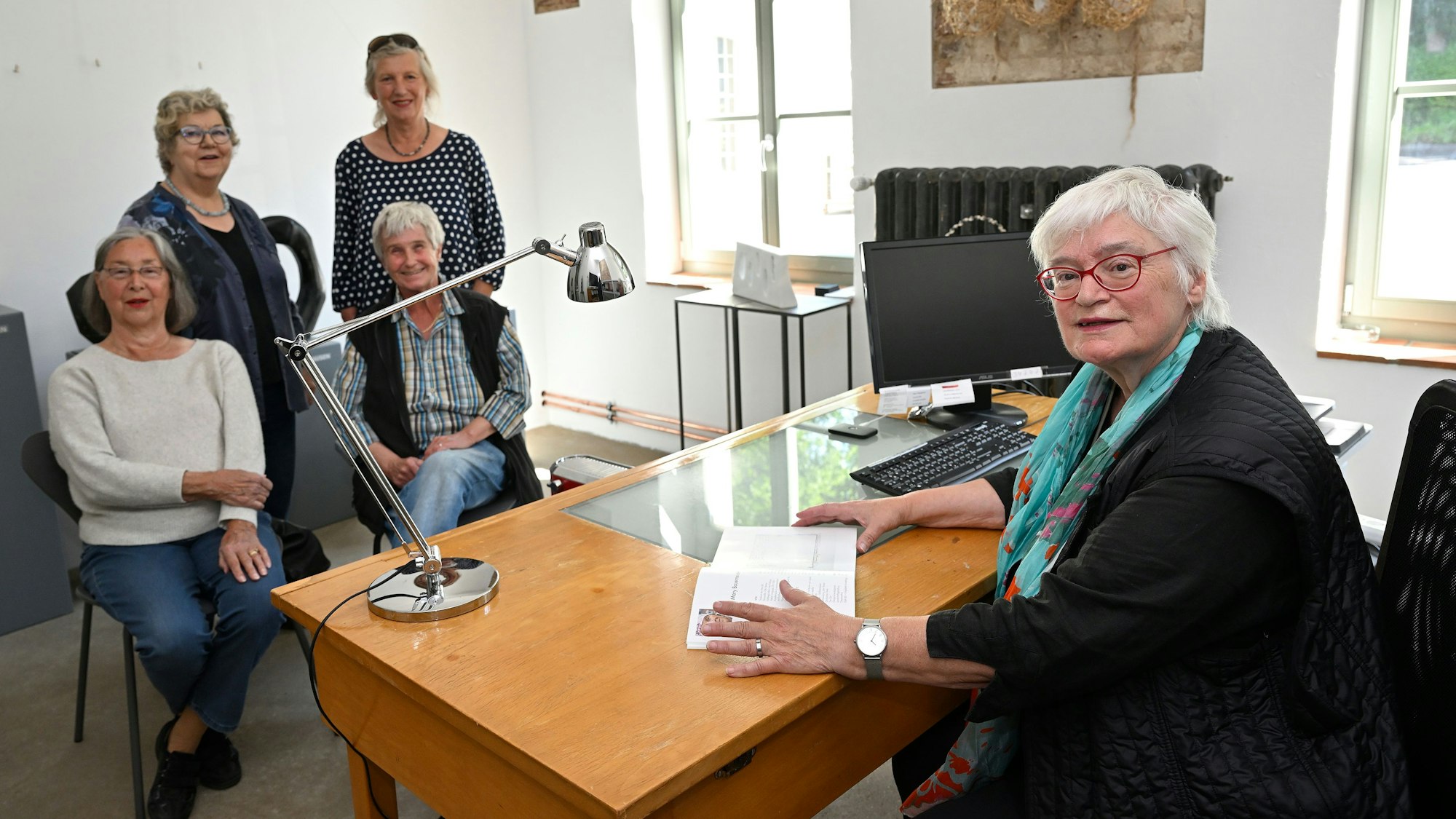 Gisela Schwarz sitzt an einem Tisch im Kunstatelier in den Räumen der Grube Weiß in Bergisch Gladbach-Moitzfeld. auf der anderen Seite des Tisches stehen und sitzen Künstlerkollegen.