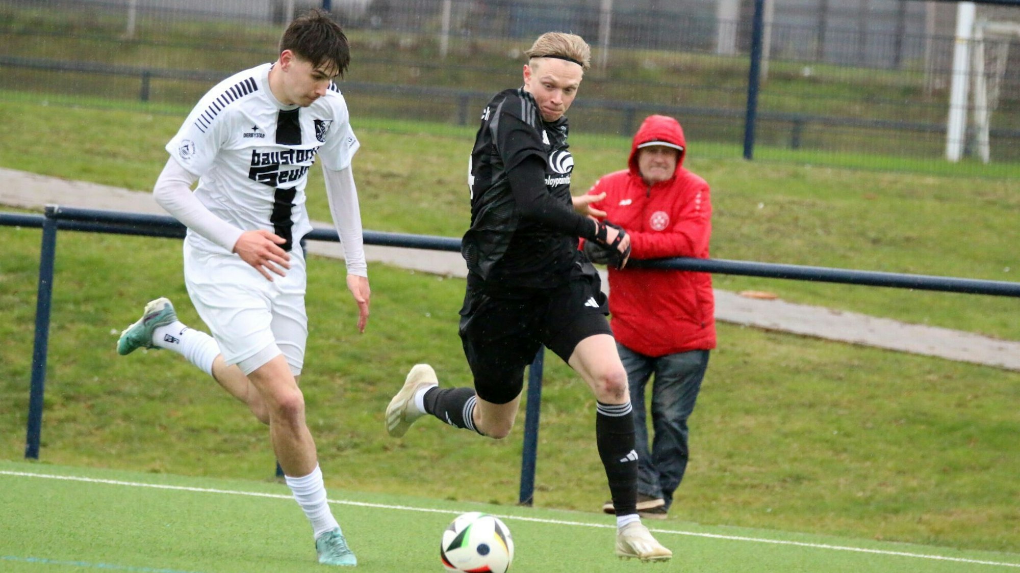 Tim Birkenheuer im Laufduell mit einem Gegenspieler. Im Hintergrund steht ein Zuschauer, der die Kapuze über den Kopf gezogen hat.