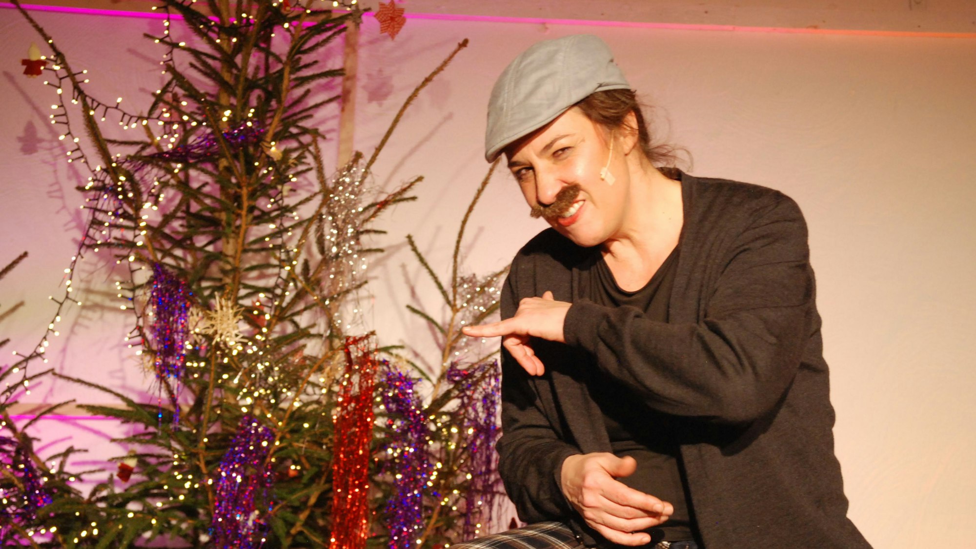 Eva Eiselt ist mit Schnurrbart und Mütze verkleidet und sitzt neben einem Weihnachtsbaum.