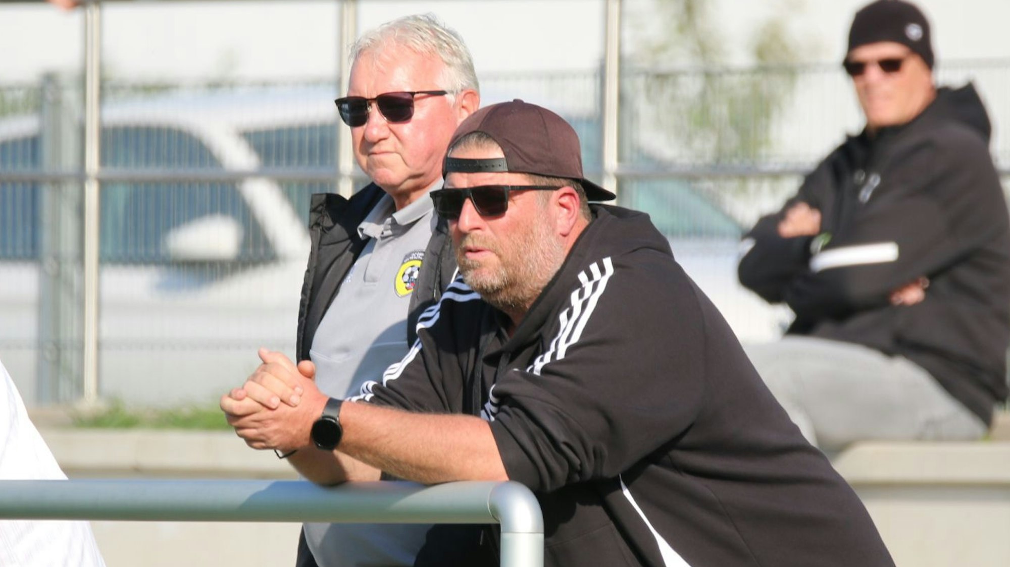 Karsten Kochems und Paul Esser stehen hinter der Reling am Sportplatz in Erftstadt. Beide tragen Sonnenbrillen.