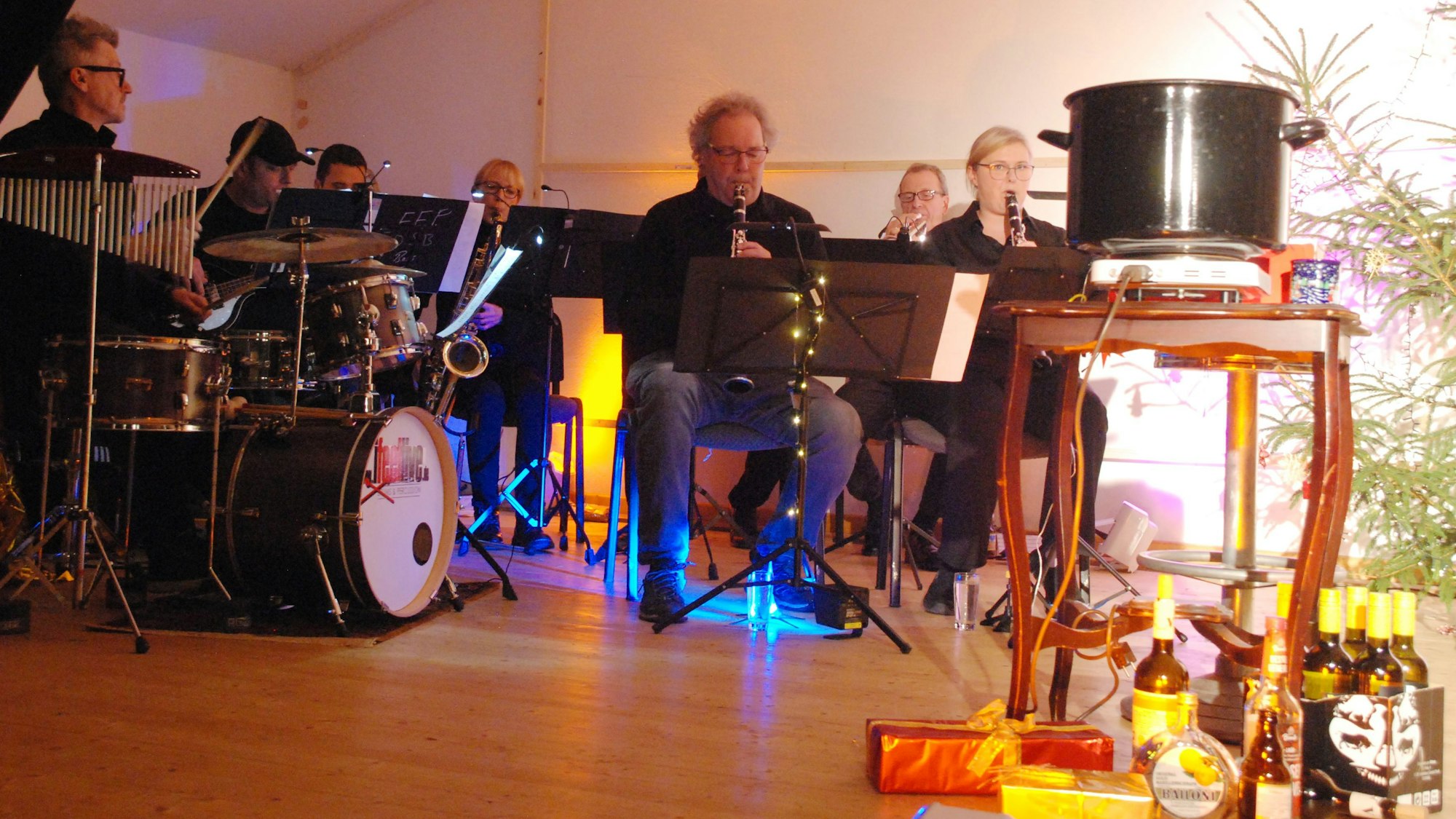 Musiker sitzen auf einer Bühne und spielen auf ihren Instrumenten. Rechts steht ein schwarzer Glühweintopf, auf dem Boden mehrere Flaschen.