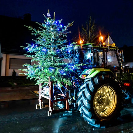 Das Bild zeigt einen Traktor, der einen beleuchteten Weihnachtsbaum transportiert.