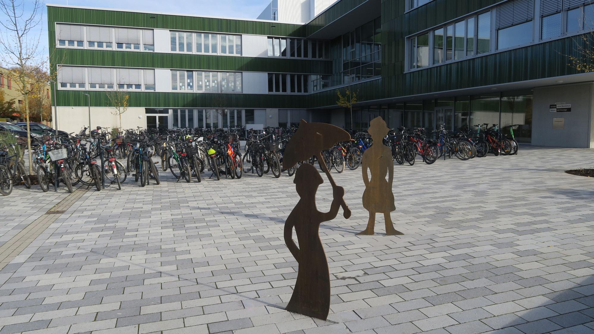 Vor einem Schulgebäude stehen viele Fahrräder und zwei Figuren.