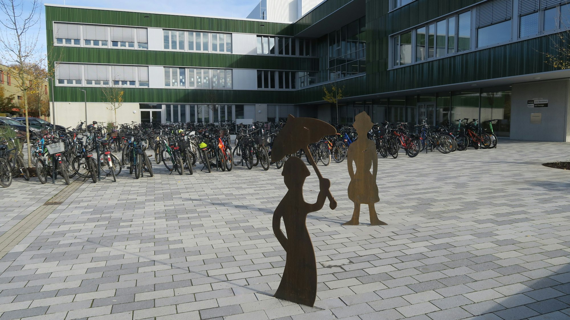 Vor einem Schulgebäude stehen viele Fahrräder und zwei Figuren.