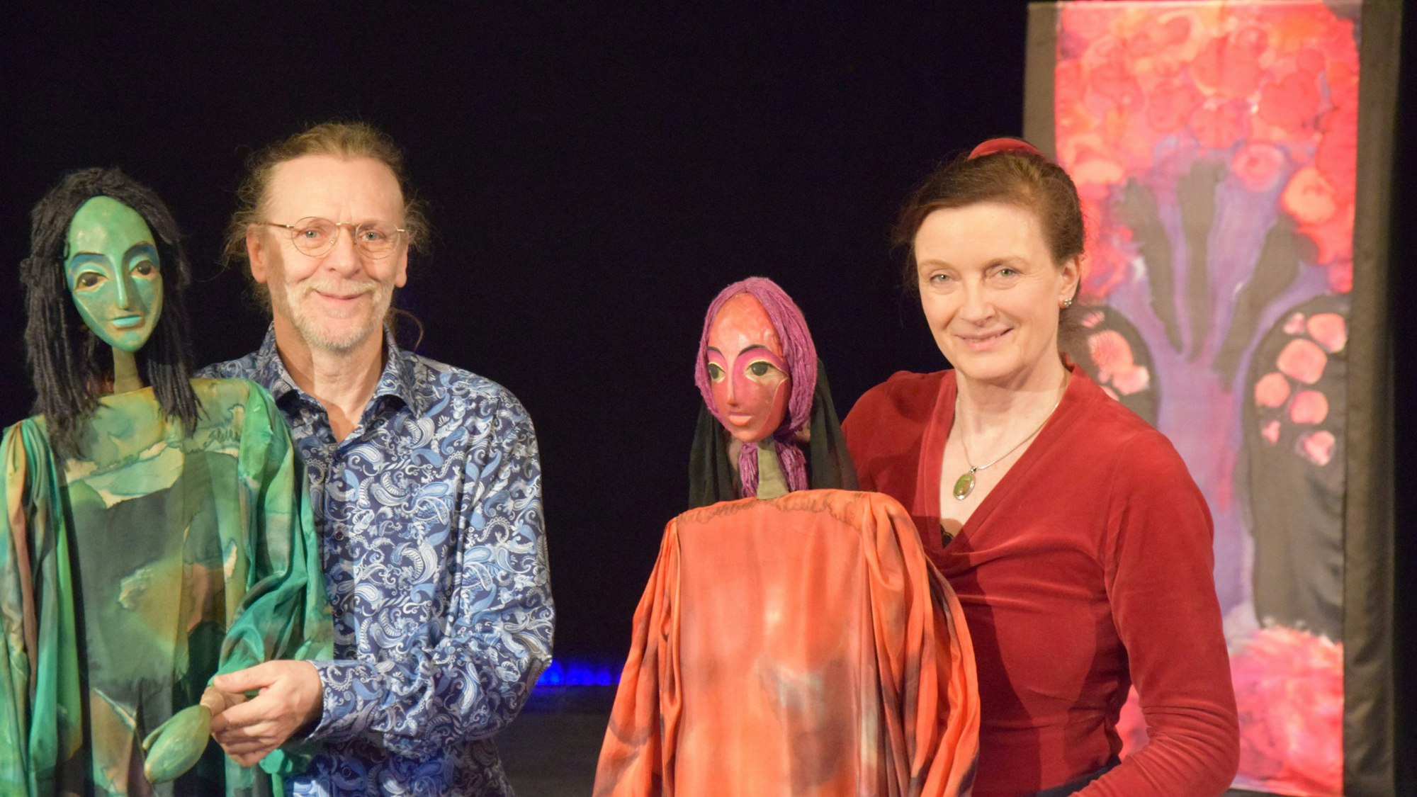 Eine Mann und eine Frau haben menschenhohe Puppen bei sich und stehen auf einer Bühne.