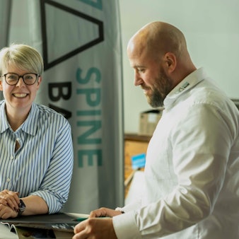 Das Bild zeigt die beiden Obengenannten an einem Stehtisch. Während Neef-Wedler in die Kamera schaut, blättert Gissinger in einem Prospekt.