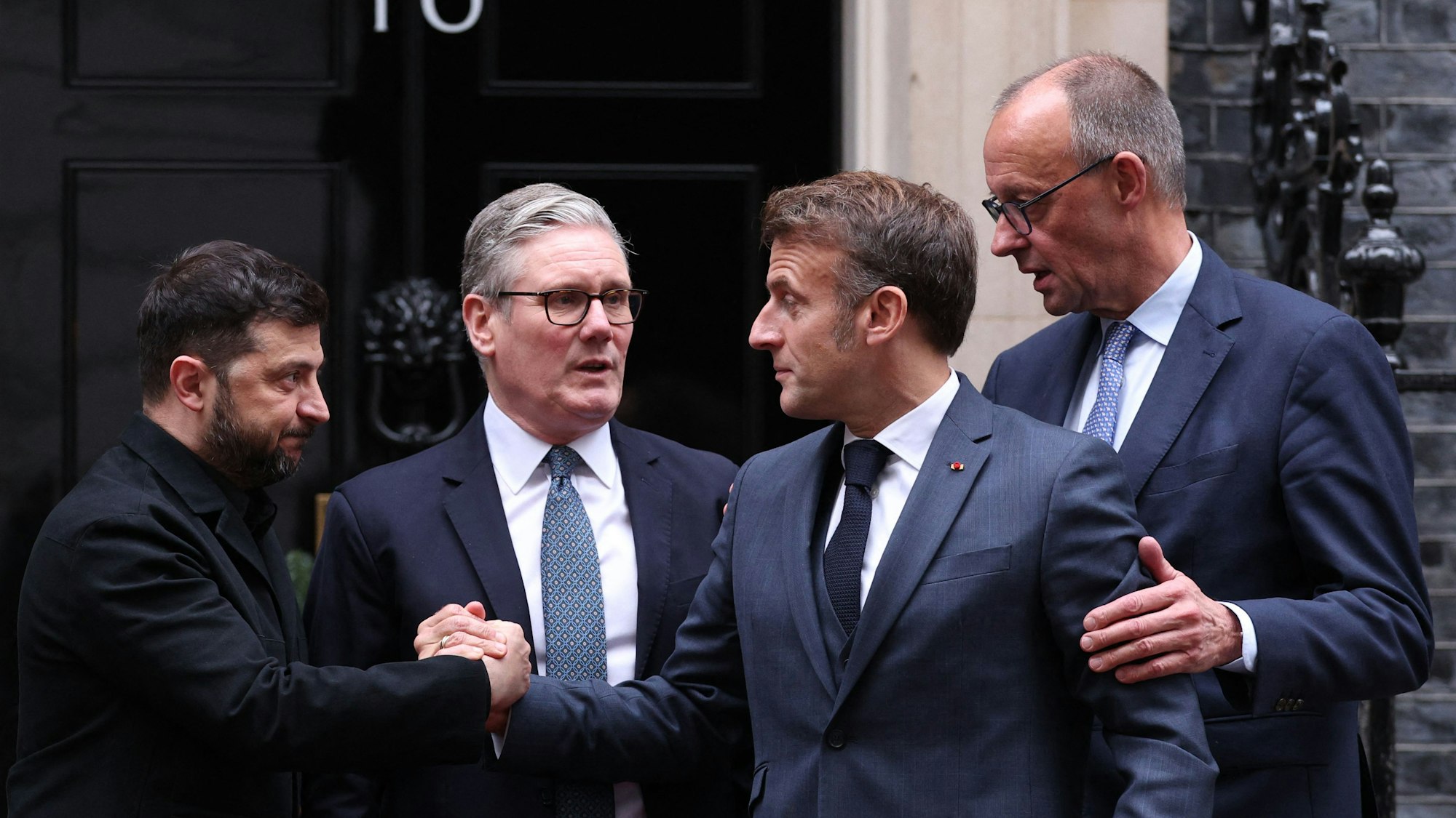 Das Bild zeigt Ukraines Präsident Wolodymyr Selenskyj (links) und Frankreichs Präsident Emmanuel Macron (3. von links) beim Händedruk vor einem Meeting in Downing Street 10 in London. Der britische Premierminister Keir Starmer (2. von links) und Bundeskanzler Friedrich Merz (ganz rechts) sind bei dem Treffen in Downing Street 10 in London auch dabei.