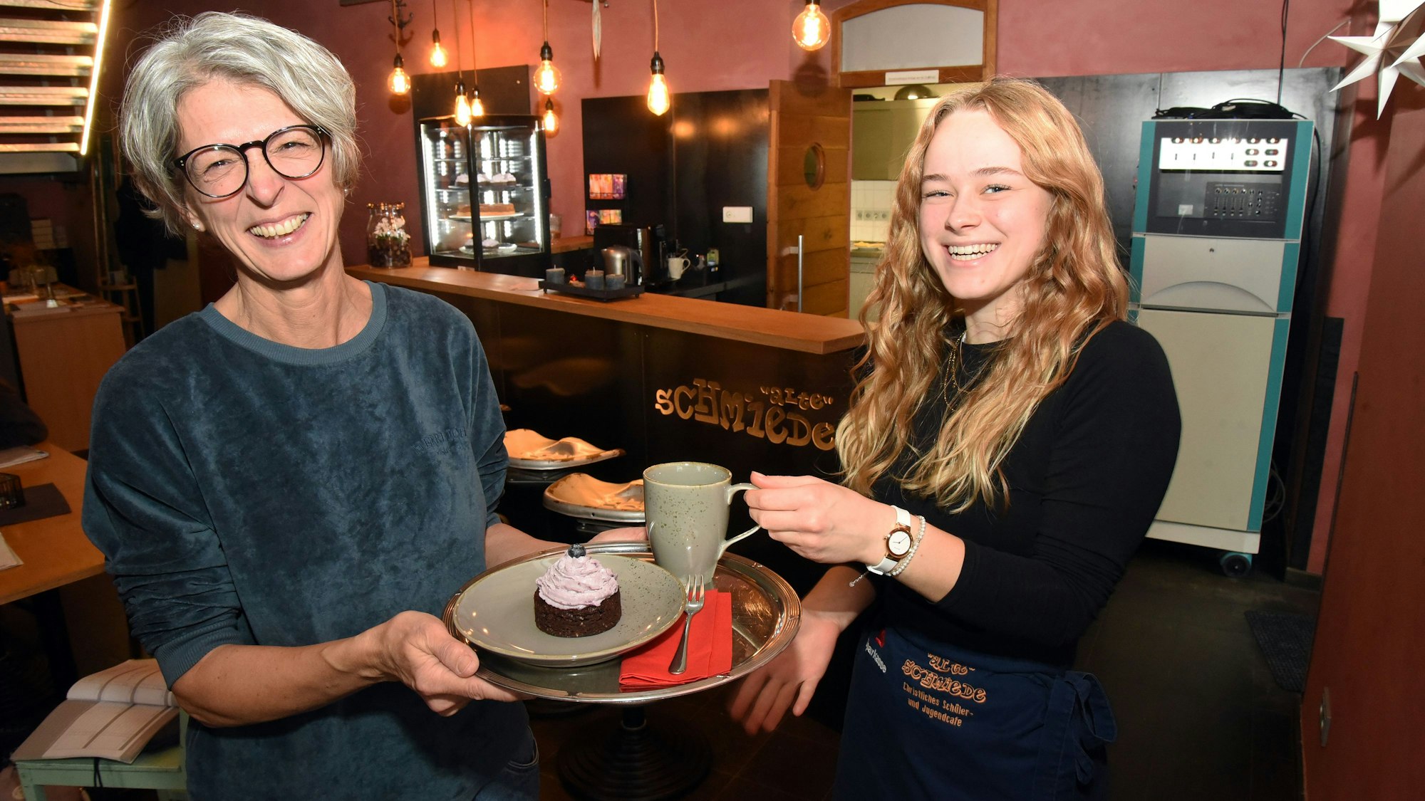 Ulrike Schuster und Bernice Schröder lachen in die Kamera, während sie einen Kaffee und ein Stück Kuchen auf einem Tablett halten.