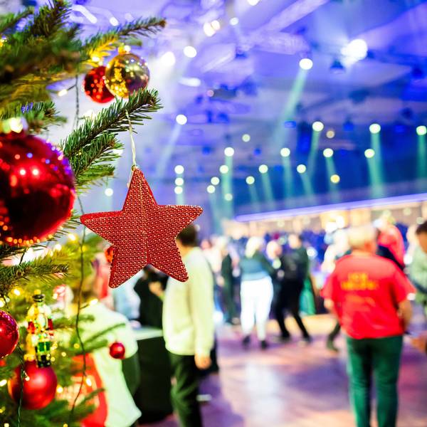 Ein geschmückter Weihnachtsbaum steht im Vordergrund. Dahinter sieht man Menschen, die in einem großen Saal feiern.