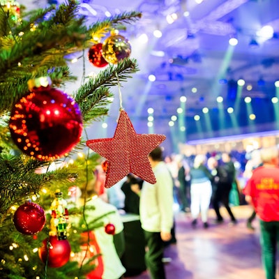 Ein geschmückter Weihnachtsbaum steht im Vordergrund. Dahinter sieht man Menschen, die in einem großen Saal feiern.