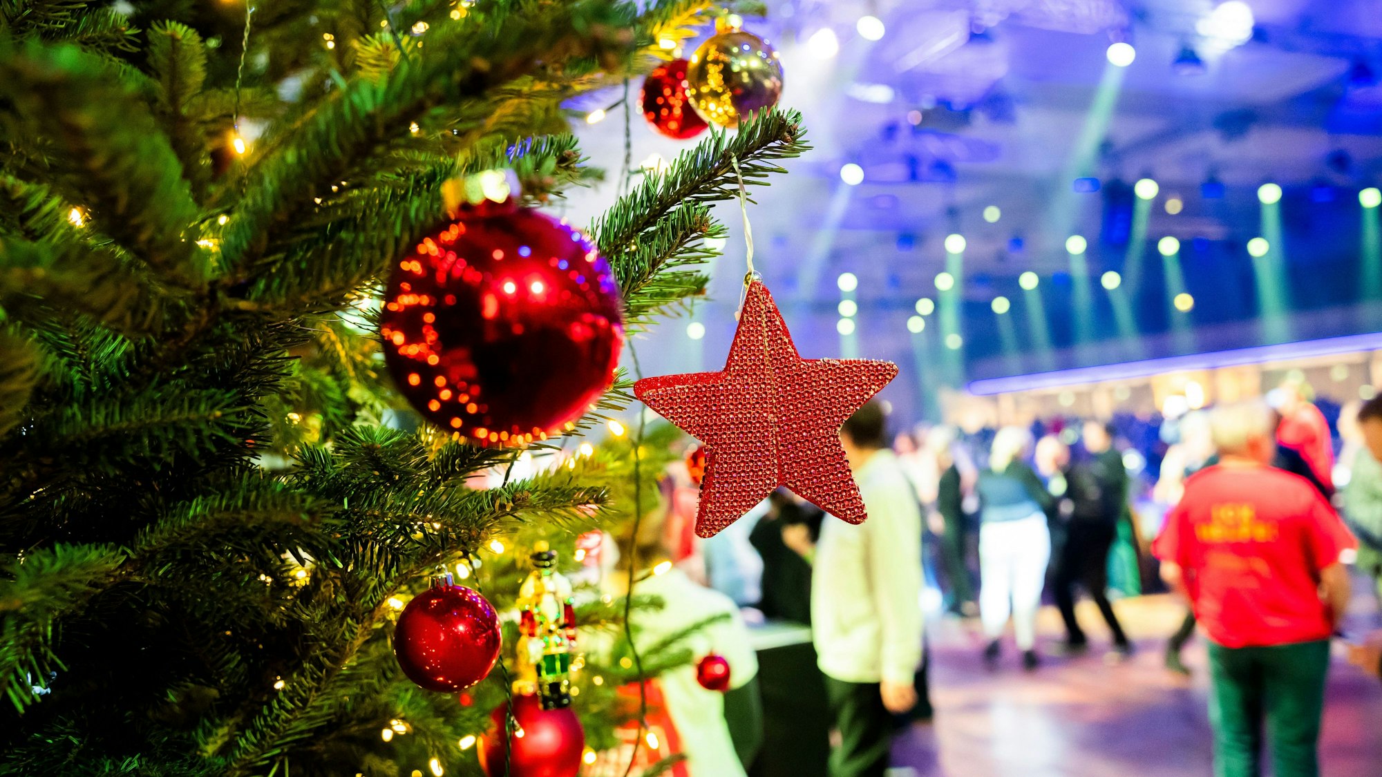 Ein geschmückter Weihnachtsbaum steht im Vordergrund. Dahinter sieht man Menschen, die in einem großen Saal feiern.