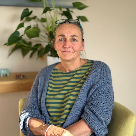 Eine Frau sitzt auf einem gelben Sessel in einem hellen Raum, hinter ihr steht eine Pflanze auf einem Regalbrett.