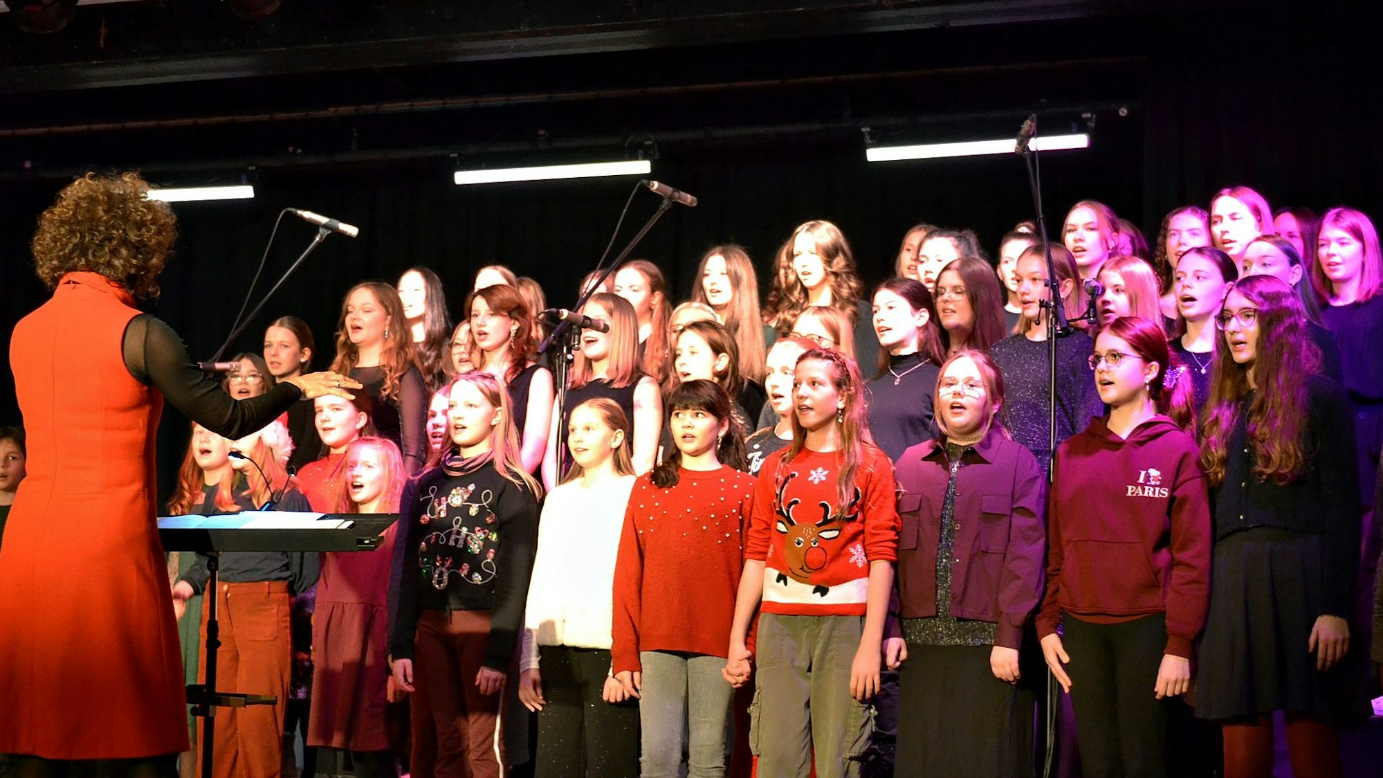 Der Chor aus Kindern und Jugendlichen steht singend auf der Bühne, alle beobachten die Dirigentin.