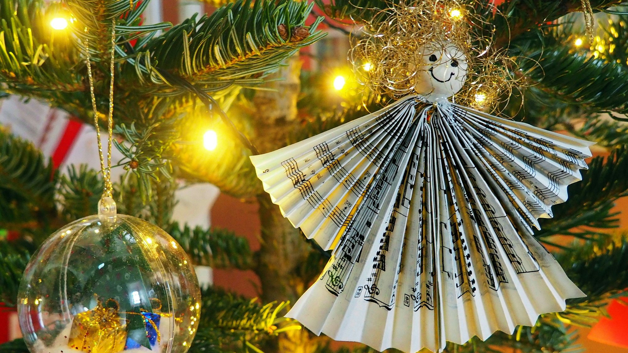 Eine Weihnachtskugel mit Watte und Geschenken darin hängt neben einem Papierengel an einem Weihnachtsbaum.