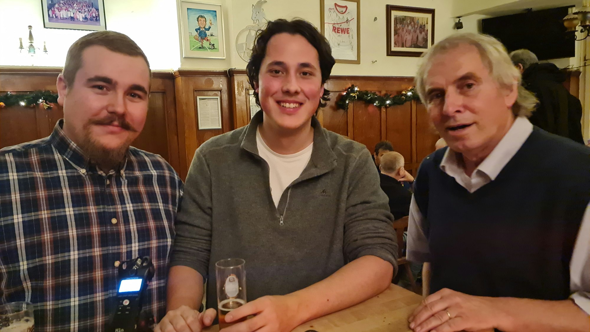 Drei Männer stehen an eine Theke, vor dem Mann ganz links steht ein Aufnahmegerät auf der Theke.