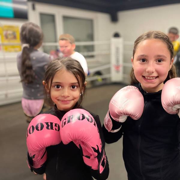 Zwei junge Mädchen stehen im Boxring des Vereins Cologne Thai Kickboxing e.V. und halten ihre rosa-farbenen Boxhandschuhe vor die Kamera.