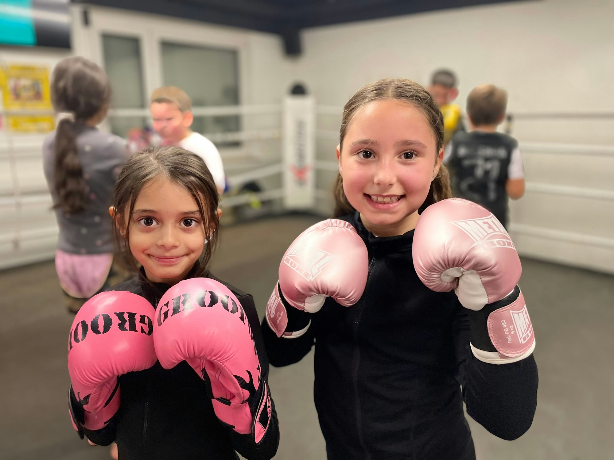Zwei junge Mädchen stehen im Boxring des Vereins Cologne Thai Kickboxing e.V. und halten ihre rosa-farbenen Boxhandschuhe vor die Kamera.