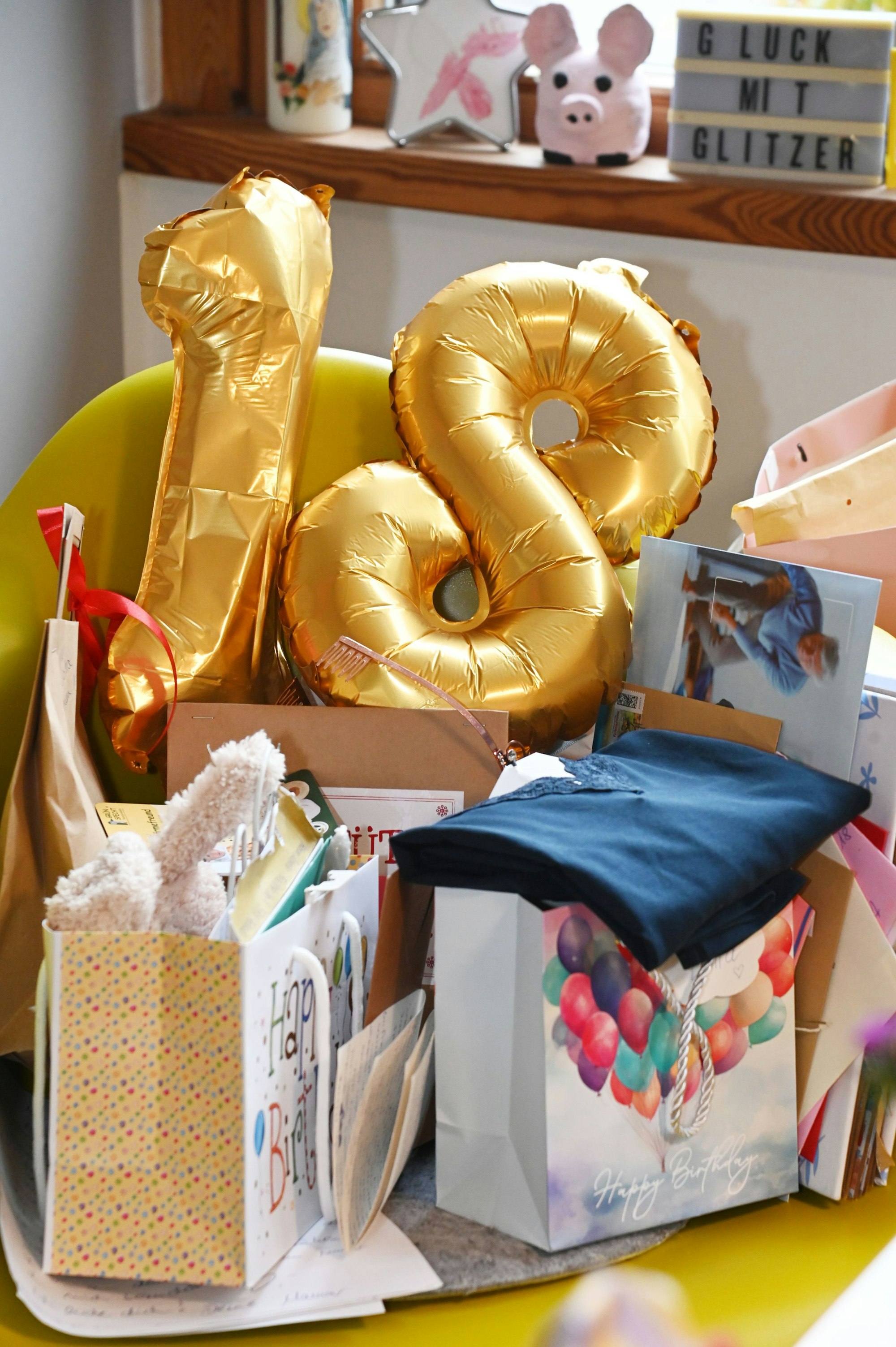 Papiertüten mit Geschenken sind zu sehen, ein goldener Luftballon formt eine 18.