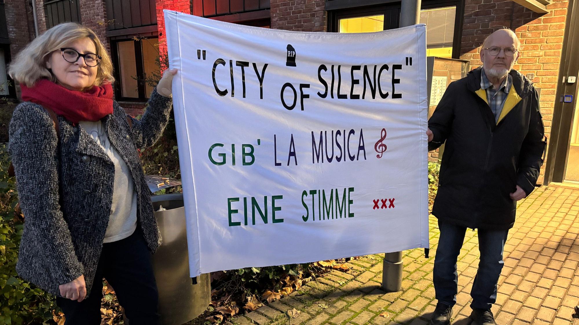 Das Foto zeigt zwei Menschen, die ein Plakat mit der Aufschrift „City of Silence Gib La Musica eine Stimme“ beschriftet ist.